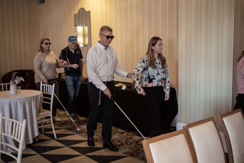 Participants being assisted while wearing low-vision glasses and walking through a conference room.